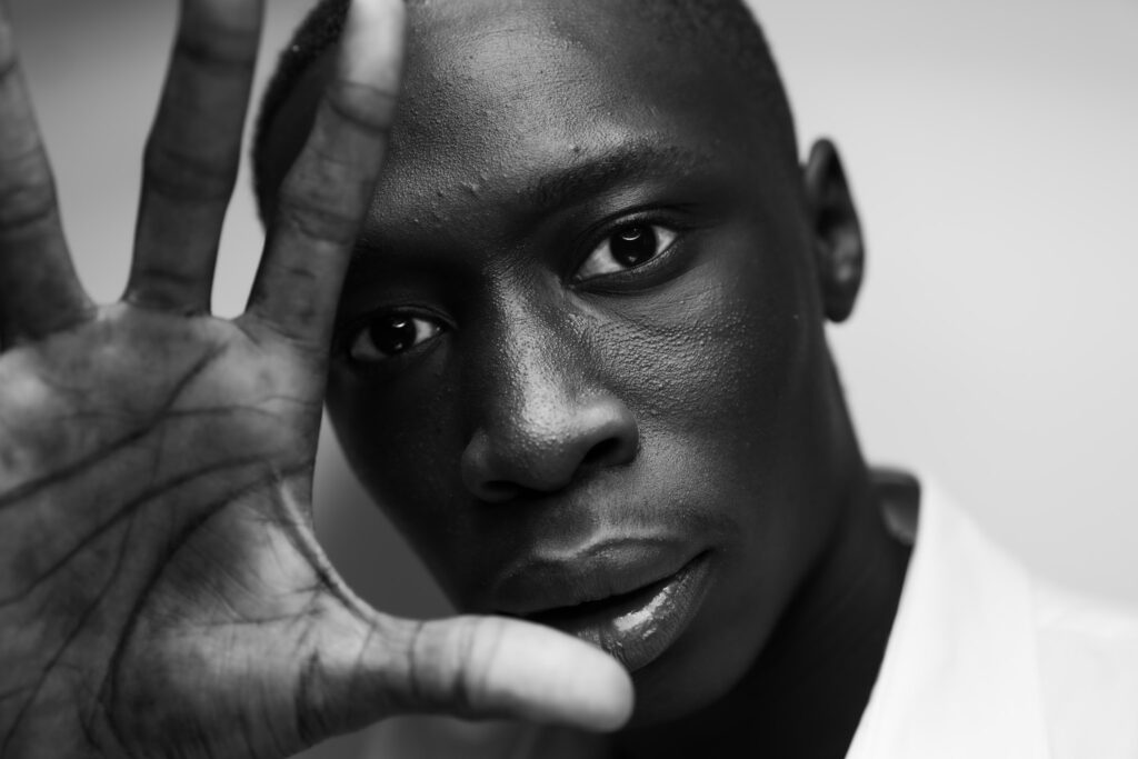 Close-up black and white portrait of Khaby Lame holding his hand open near his face in his signature gesture, looking directly at the camera with a neutral expression.